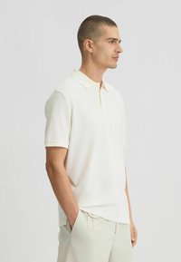 Polo di colore avorio, maniche corte, colletto classico, tessuto morbido. Abbinato a pantaloni chiari, che mostrano un fit e uno stile rilassati.