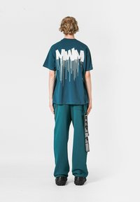 Camiseta de algodón verde azulado con gráfico blanco goteando en la parte de atrás, combinada con pantalones verdes azules con bolsillos laterales y etiquetas, y zapatos negros.