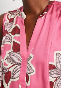 Ärmlös blus i rosa tyg med ett blommönster som har röda och vita blad, accentuerad av en knäppt krage och v-ringning.