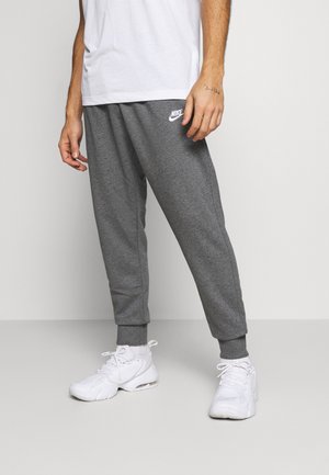 Mann trägt graue Nike-Jogginghose, weiße Socken und weiße Turnschuhe, steht vor einem schlichten hellen Hintergrund.