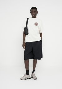 Λευκό υπερμεγέθες t-shirt με καφέ εκτύπωση, μαύρο σορτς και γκρι ανοιχτά αθλητικά παπούτσια. Μικρή μαύρη τσάντα φορεμένη χιαστί. Λείες υφές σε όλο το σύνολο.