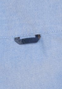 Blauwe stof met een subtiele textuur, met een zwarte plastic clip die in het materiaal is verwerkt aan de rand, nabij de stikdetails.