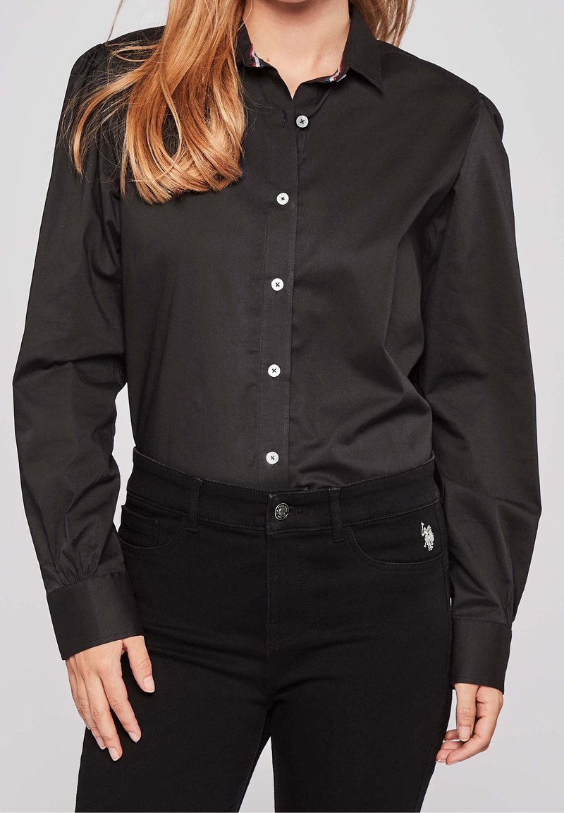 Chemise noire boutonnée avec col, manches longues et boutons blancs. Le tissu semble lisse avec une légère brillance ; portée avec un jean noir.
