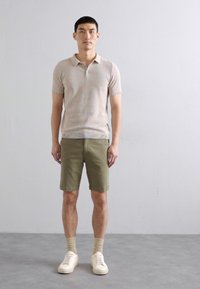 Beige rövid ujjú polo póló, olajzöld rövidnadrág, fehér sportcipő és bézs zokni. A modell egy sima világosszürke háttér előtt áll.