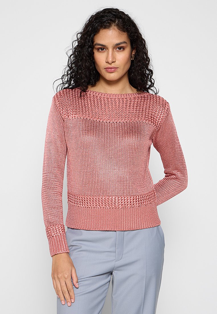 Donna con capelli ricci, che indossa un maglione lavorato a maglia rosa polveroso e pantaloni grigi chiari, in posa con una mano dietro la schiena.