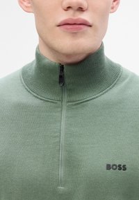 Green knit pullover with a high collar, half-zip closure, and ribbed accents. Features a black logo on the left chest.