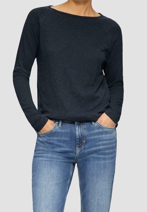 Persona con camiseta de manga larga azul marino oscura metida dentro de unos vaqueros desgastados de color azul claro, con las manos en los bolsillos, contra un fondo liso.