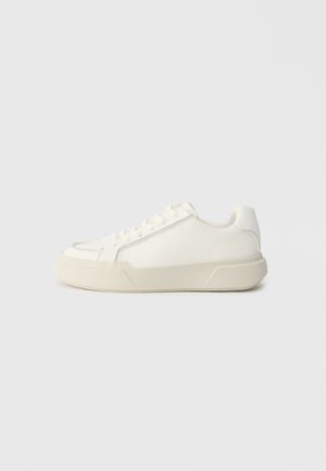 White sneaker made of smooth leather, featuring a rounded toe, lace-up front, and a thick rubber sole with an angular design.