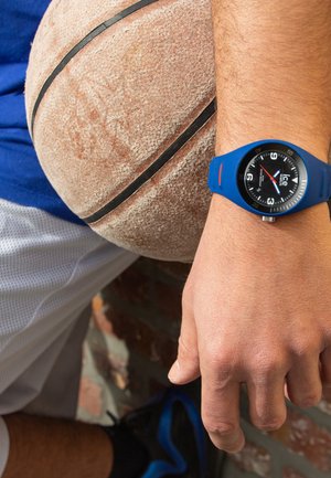Blue sports watch with a black dial, white markings, and a red second hand, worn on a hand resting against a textured basketball.