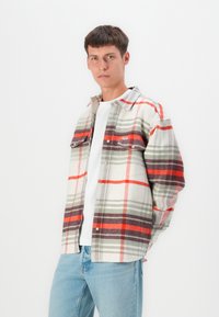 Camisa de cuadros hecha de una tela suave, con rayas rojas, grises y blancas. Cuello clásico, dos bolsillos en el pecho y botones a presión. Lleva puesta una camiseta blanca debajo.