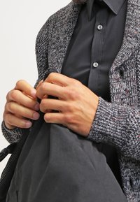 Graue gestrickte Strickjacke mit strukturierten Mustern, schwarzes Hemd darunter und Hände, die eine schwarze Tasche halten, wobei ein Knopfdetail am Hemd zu sehen ist.