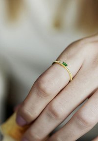 Anello d'oro con una fascia sottile, caratterizzato da una pietra verde rettangolare e piccole pietre trasparenti su un lato. Texture liscia e design delicato.