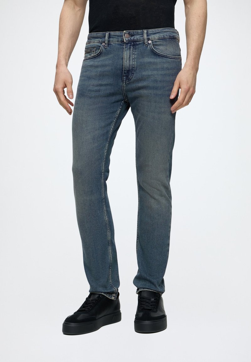 Mann trägt ausgewaschene blaue Jeans und schwarze Turnschuhe und steht vor einem schlichten weißen Hintergrund.