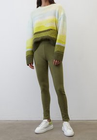 Den stickade tröjan har horisontella ränder i grönt, gult och vitt; den är kombinerad med olivgröna leggings och vita sneakers med gröna detaljer.