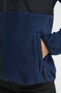 Veste en polaire marine avec partie supérieure noire, poche latérale zippée et poignet élastique. Texture épaisse et douce dans un design à coupe décontractée.