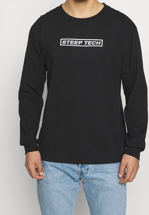 T-shirt noir à manches longues présentant un logo rectangulaire blanc avec le texte "STEEP TECH" sur la poitrine ; en coton, coupe décontractée.