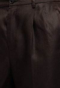 Pantalon marron foncé sur mesure avec plis à l'avant, poches latérales et fermeture par bouton à la taille.