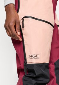 Červené a ružové nepremokavé zateplené nohavice s čiernym zipsom, s dvojtónovým dizajnom a vyšitým nápisom "R&D" na vrecku.