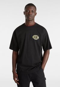 Camiseta de manga corta negra con un logo ovalado amarillo de "VANS" en el pecho, hecha de algodón suave, con un corte relajado y cuello redondo.