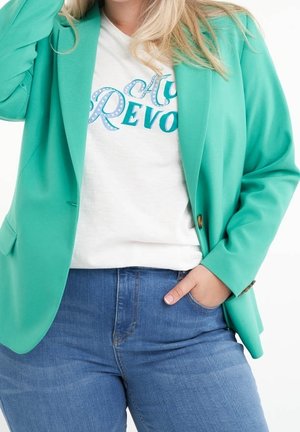 Vrouw draagt een muntgroene blazer over een wit shirt met opdruk en blauwe jeans, met één hand in de spijkerbroekzak, tegen een witte achtergrond.