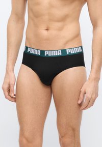 Αρσενικός κορμός που φοράει μαύρα εσώρουχα με πράσινη μέση που φέρει λευκό κείμενο "PUMA", στέκεται μπροστά σε απλό φωτεινό φόντο.