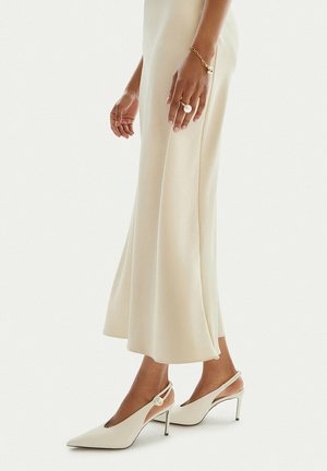 Frau trägt ein elfenbeinfarbenes Satin-Midikleid, spitze Slingback-Absatzschuhe mit Perlendetail, ein goldenes Armband und einen großen Perlenring an der linken Hand.