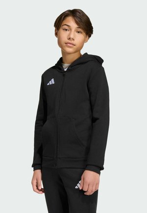 Giovane persona che indossa una felpa nera con zip e pantaloni abbinati con il logo Adidas bianco, in piedi davanti a uno sfondo chiaro.