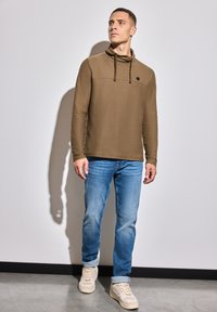 Hnědý svetr s vysokým límcem a šňůrkami, kombinovaný s světle modrými džíny. Na hrudi se nachází černý akcent.