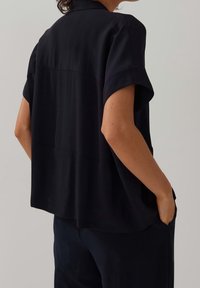 Top corto nero a maniche corte in un tessuto morbido e liscio, con una vestibilità comoda, caratterizzato da un yoke posteriore e tasche laterali per una funzionalità aggiuntiva.