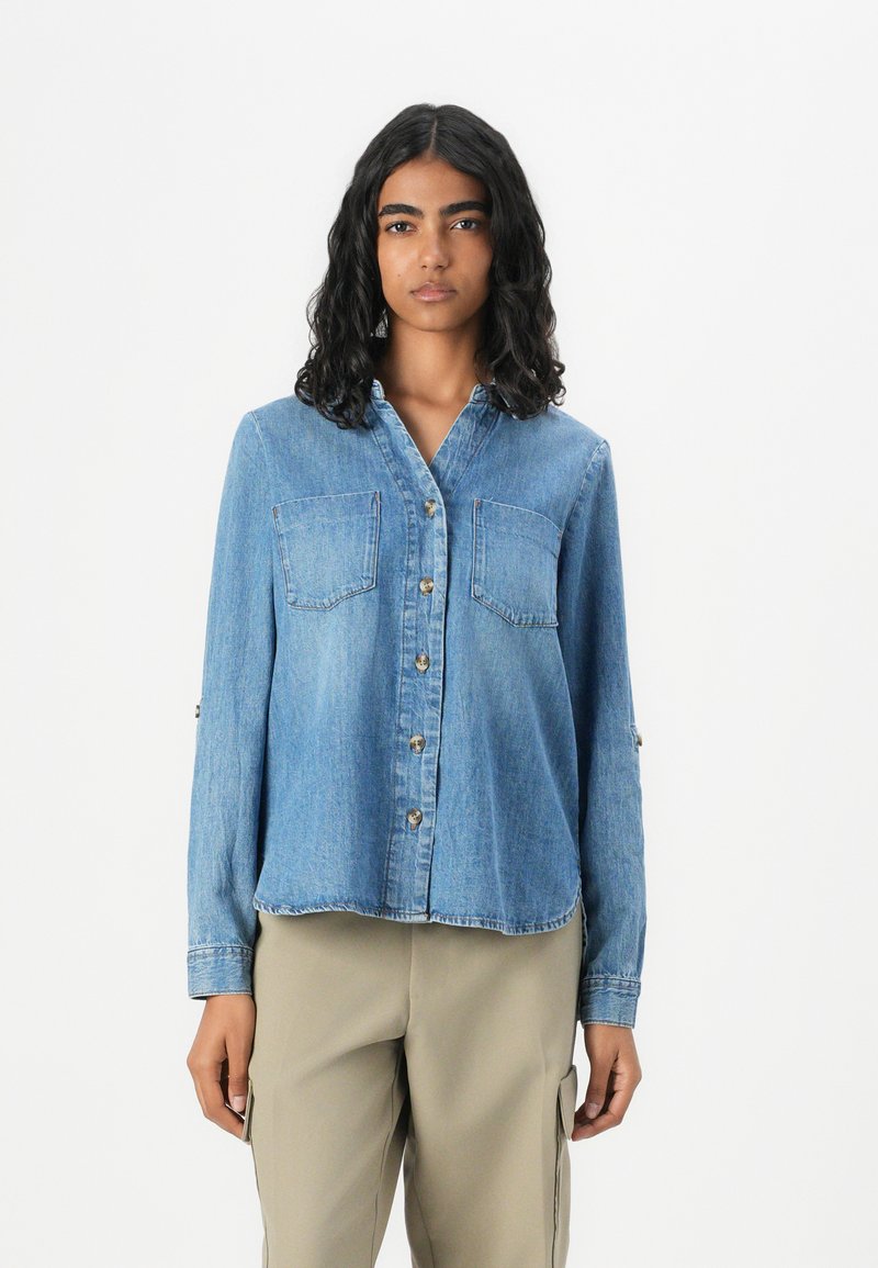 Light blue denim shirt with a collar, front button closure, and two chest pockets. Features long sleeves with button details.
