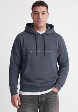 Homme portant un sweat à capuche Jack & Jones bleu foncé avec une poche avant, un t-shirt blanc en dessous, et un jean gris, debout devant un fond blanc.