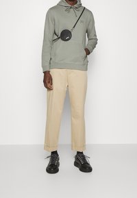 Sudadera con capucha de color verde claro con cordón, pantalones beige de pierna ancha y zapatillas negras con cordones estampados, complementados con un bolso redondo negro.