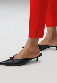 Zwarte leren mules met spitse neuzen en een kleine hak, met een strikaccent op de wreef en gedragen met rode cropped broek.