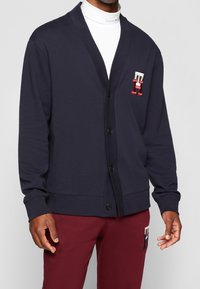 Cardigan blu navy realizzato in tessuto morbido, con scollo a V, chiusura con bottoni e toppa con logo in rosso e bianco. Indossato sopra un dolcevita bianco.