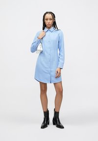 Lichtblauwe hemdjurk met een knoopsluiting aan de voorkant, kraag en lange mouwen; gecombineerd met zwarte enkellaarsjes. Glad stoffen textuur, eenvoudig ontwerp.