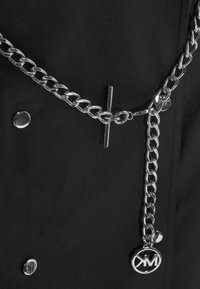 Ceinture chaîne en argent avec fermoir bascule et pendentif rond gravé "KM" sur un fond en tissu noir avec des boutons argentés.