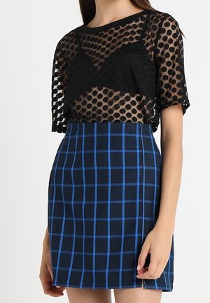 Femme portant un haut transparent noir à pois sur un soutien-gorge noir, associé à une jupe taille haute bleu marine avec un motif à carreaux bleus.