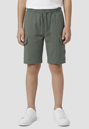 Enfant portant un short cargo vert olive avec des poches avant et des baskets blanches, debout devant un fond uni.