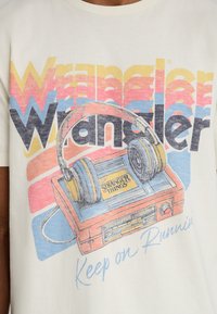 Cream t-shirt featuring a retro cassette player illustration with headphones, and multicolored "Wrangler" text; includes "Stranger Things" logo.