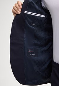 Interno di un giubbotto in lana stretch blu con foderatura liscia, una piccola tasca con dettagli bianchi e etichette del marchio.