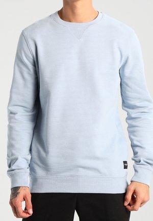 Mężczyzna ubrany w jasnoniebieską bluzę typu crewneck z prążkowanymi mankietami i dołem, w połączeniu z czarnymi spodniami, stojący na jednolitym tle.