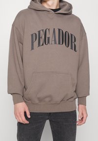 Felpa con cappuccio marrone con la scritta "PEGADOR" in nero disposta ad arco. Presenta una tasca frontale e polsini a coste, realizzata in tessuto morbido e accogliente.