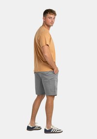 Camiseta de algodón marrón y pantalones cortos de carga grises. Los pantalones cortos tienen bolsillos laterales. Se llevan con zapatillas blancas con detalles en azul y marrón.