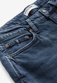 Jean boyfriend bleu foncé décontracté avec poche avant, rivets en cuivre, passant de ceinture, bouton et étiquette visible à l’intérieur de la ceinture.