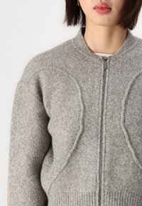 Sivi zip-up pulover napravljen od meke vune, s rebrastim rubom i jedinstvenim teksturiranim detaljima na ramenima. Glatka površina i okrugli izrez.