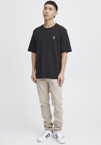 Black cotton t-shirt with a graphic detail on the left chest, paired with beige trousers, and white sneakers featuring black stripes.