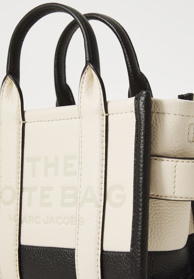 Small tote bag featuring a cream-colored leather body with black accents, top handles, and embossed text reading "THE TOTE BAG."