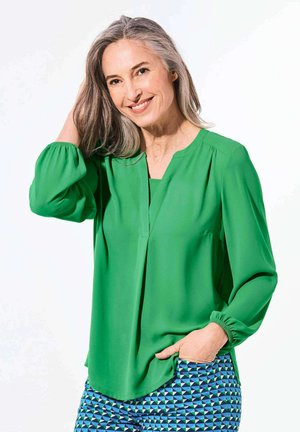 Blusa verde con scollo a V, dettagli a pieghe e maniche lunghe a sbuffo. Abbinata a pantaloni blu e verdi con motivi geometrici.