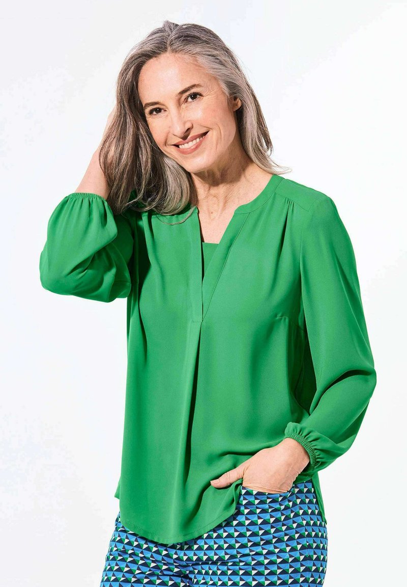 Grüne Bluse mit einem V-Ausschnitt, Faltendetails und langen Puffärmeln. Kombiniert mit gemusterten blau-grünen Hosen, die geometrische Formen aufweisen.