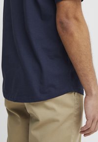 Tricou din bumbac de culoare bleumarin, expus dintr-un unghi, cu guler rotund și mâneci scurte, asortat cu pantaloni din bumbac bej.